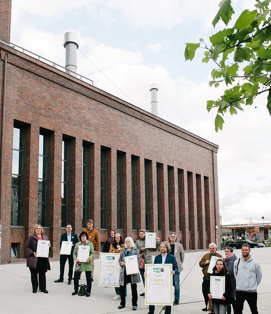Akteur:innen des Aktionsbündnis Fairer Handel in Berlin bei der Wiederauszeichnung als Fairtrade Town halten ihre Titel in die Kamera.