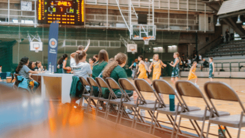 In einer Basketballhalle spielen junge Spielerinnen im Hintergrund gegeneinander, vorne sitzen Personen auf der Bank.